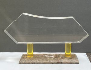 Carved clear abstract shaped plexiglass mounted on two yellow clear plexiglass poles and posted on a rectangular marble base