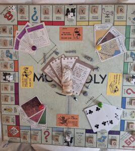 mixed media – assemblage piece on a monopoly board using other game pieces, chess, clue, scrabble dice and more to create a cohesive, fun work of art