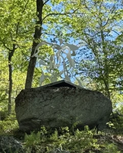Utilizing electronic cutting technology and painted stainless steel, the sculpture transforms rigid metal into fluid, pristine white ribbons that intertwine atop a rugged forest boulder, creating a powerful dialogue between industrial art and nature that symbolizes the enduring vitality of the human journey.