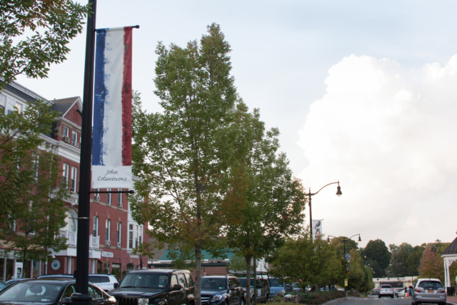 John Colantuono's red, white and blue striped banner with view of street, trees and the brick dutchess house