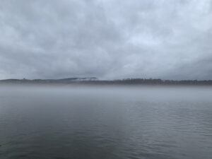 Grey cloudy sky reflects in moving river flow topped with fog bisected by a distant horizon of tree tops