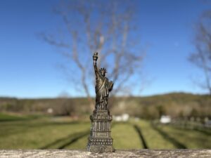 selective focus on statue of liberty sitting on weathered fence soft focus on background of meadow, large trees and distant trees