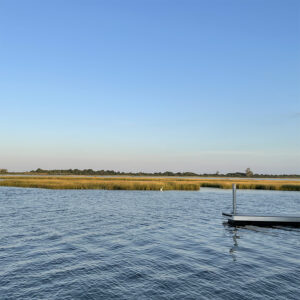 cloudless blue sky reflected in moving bay waters bisected by strip of marsh and distant tree tops