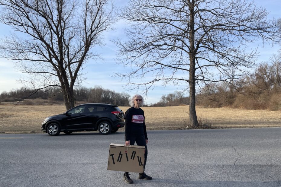 Woman standing on road, holding cardboard suitcase with test THINK behind here a car drives by and two trees frame a field