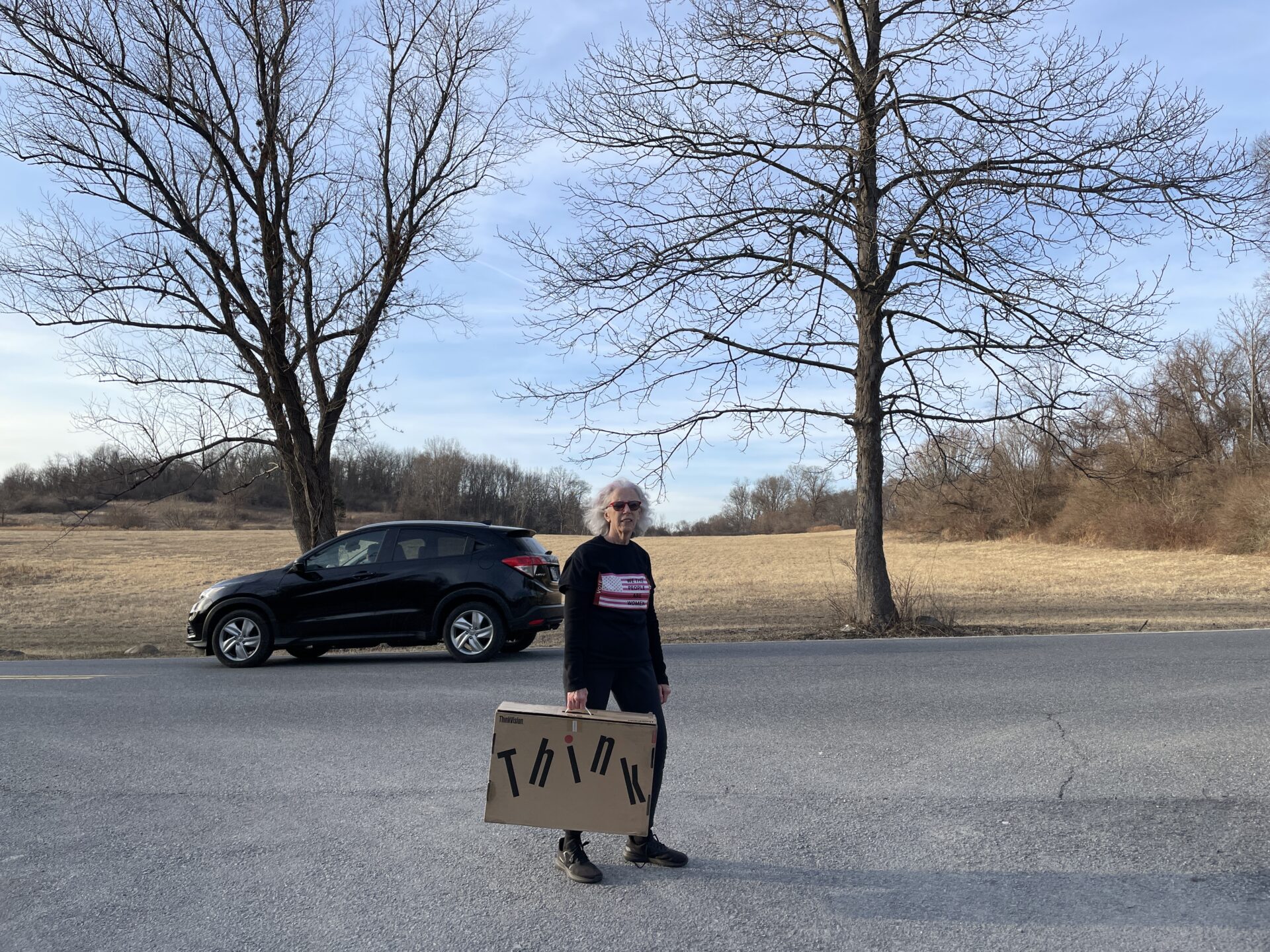 Woman standing on road, holding cardboard suitcase with test THINK behind here a car drives by and two trees frame a field