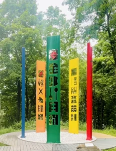 Through five monumental, vibrantly colored steel columns adorned with intricate geometric cutouts and cultural icons, artist transforms industrial metal into a dynamic visual narrative that gracefully embodies the communal spirit of cultural celebrations.