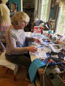 fabric artist sits by windows at table that that is covered with tools and pieces of fabric